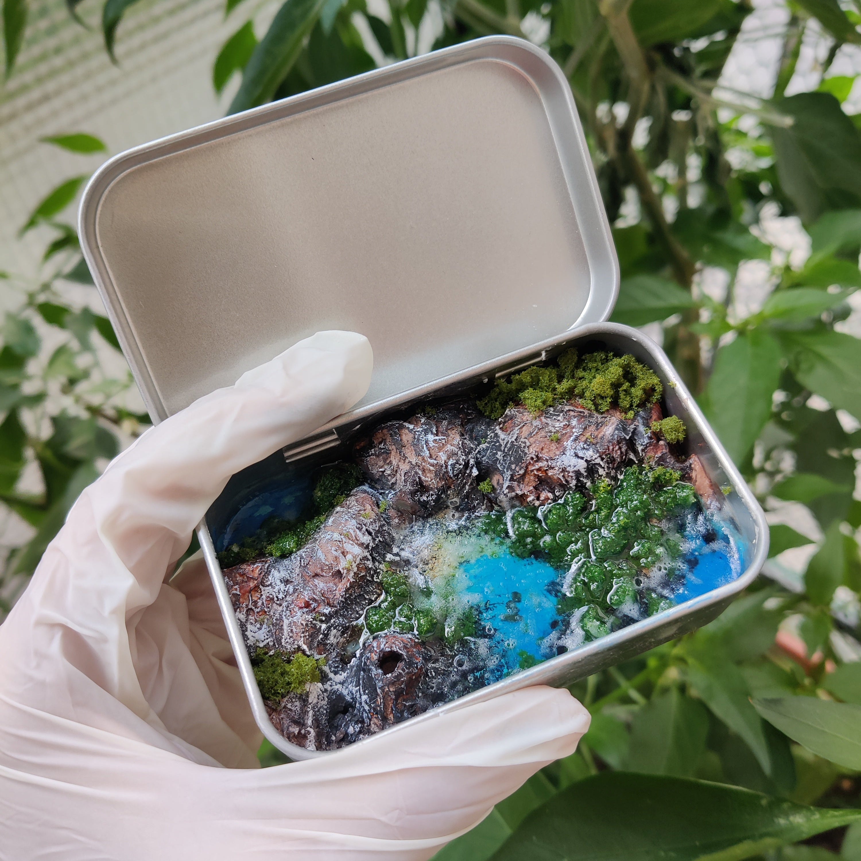Dettaglio di un diorama artigianale con scogliera, vegetazione verde e mare in resina azzurra.